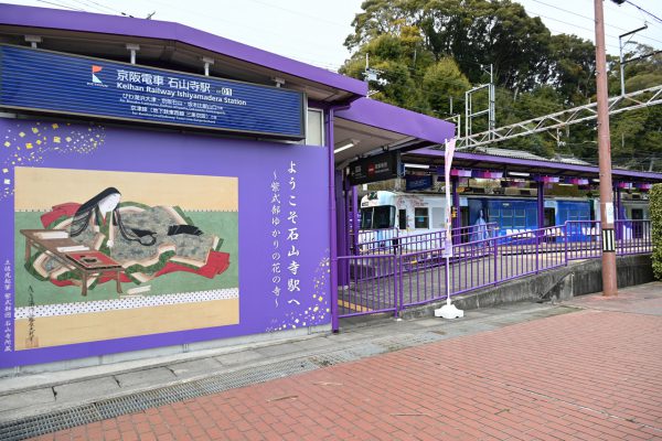 「紫式部図」をプリントした看板を設置した京阪電車石山寺駅とラッピング電車